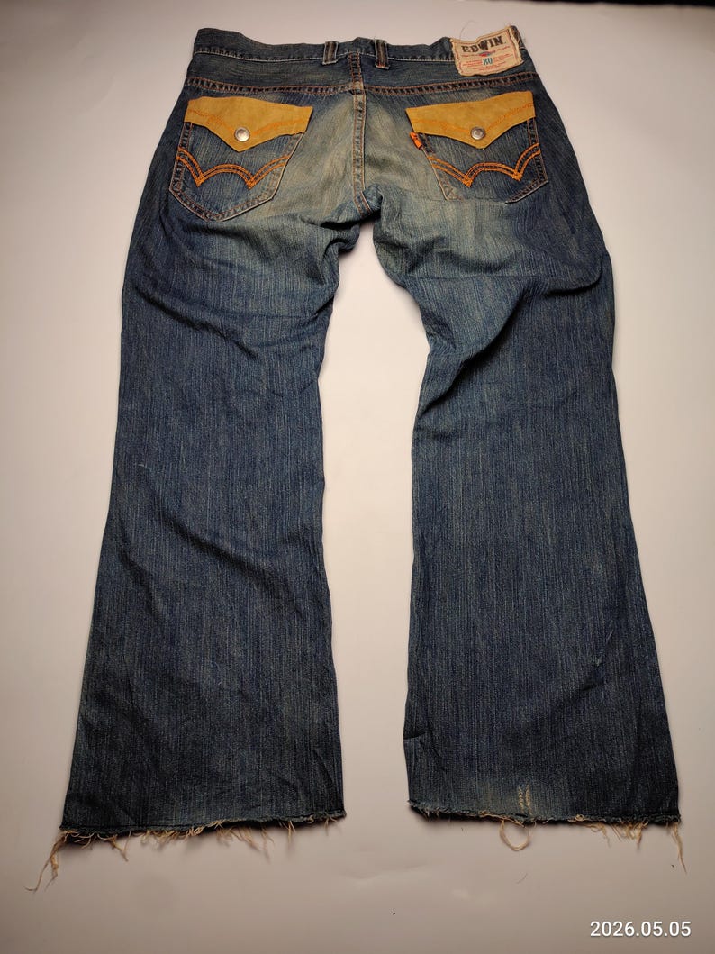 May include: Blue denim jeans with light brown suede-like back pockets and orange stitching. The jeans have a faded wash and a wide leg. The Edwin brand label is visible on the back. The jeans are laid flat on a white surface.