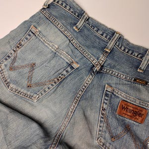 May include: Close-up of light blue denim jeans with the Wrangler logo on a brown leather patch. The jeans have classic five-pocket styling, with the signature "W" stitching on the back pockets. The jeans are slightly faded, giving them a worn-in look.
