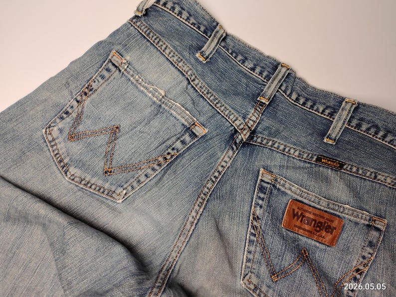 May include: Close-up of light blue denim jeans with the Wrangler logo on a brown leather patch. The jeans have classic five-pocket styling, with the signature "W" stitching on the back pockets. The jeans are slightly faded, giving them a worn-in look.