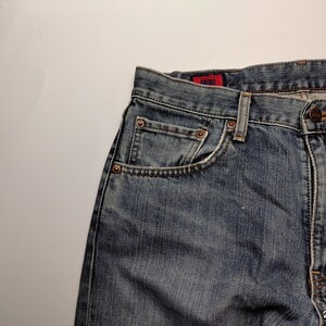 May include: Close-up of blue denim jeans with a faded wash. The jeans feature a classic five-pocket design, copper rivets, and a button closure. The red tag is visible on the back pocket. The jeans are made of durable denim.