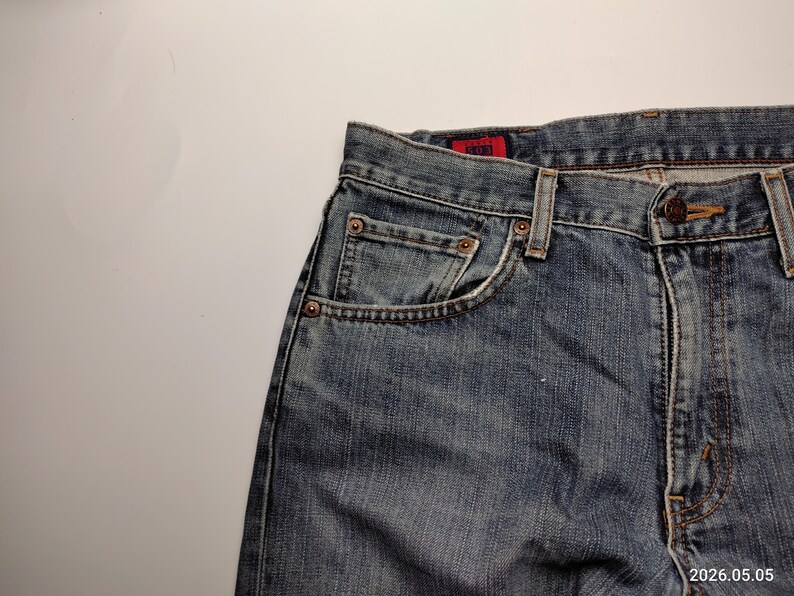 May include: Close-up of blue denim jeans with a faded wash. The jeans feature a classic five-pocket design, copper rivets, and a button closure. The red tag is visible on the back pocket. The jeans are made of durable denim.