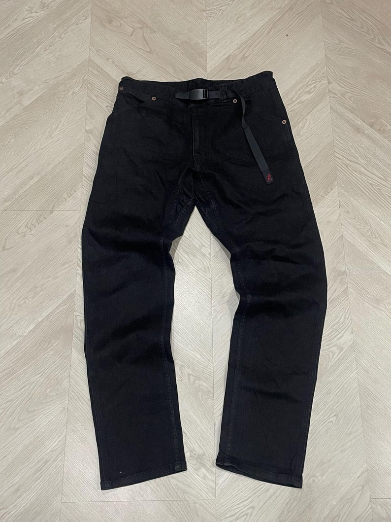 Puede incluir: Pantalones vaqueros negros con cintur&oacute;n negro incorporado. Los pantalones tienen un dise&ntilde;o de pierna recta y un detalle rojo en la trabilla del cintur&oacute;n. Se encuentran planos sobre una superficie clara con vetas de madera.