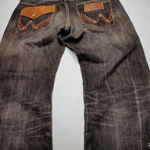 May include: Dark wash denim jeans with brown leather pocket accents and decorative stitching in orange and yellow. The jeans have a classic five-pocket design and a relaxed fit.