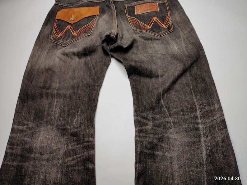 May include: Dark wash denim jeans with brown leather pocket accents and decorative stitching in orange and yellow. The jeans have a classic five-pocket design and a relaxed fit.