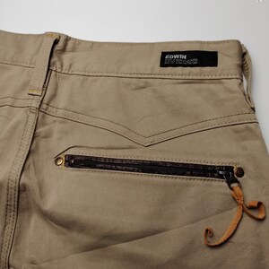 May include: Close-up of khaki-colored pants with a zipper pocket and a leather pull tab. The pants have a black label that reads "EDWIN XV KHAKI". The stitching is visible, and the overall design is casual.