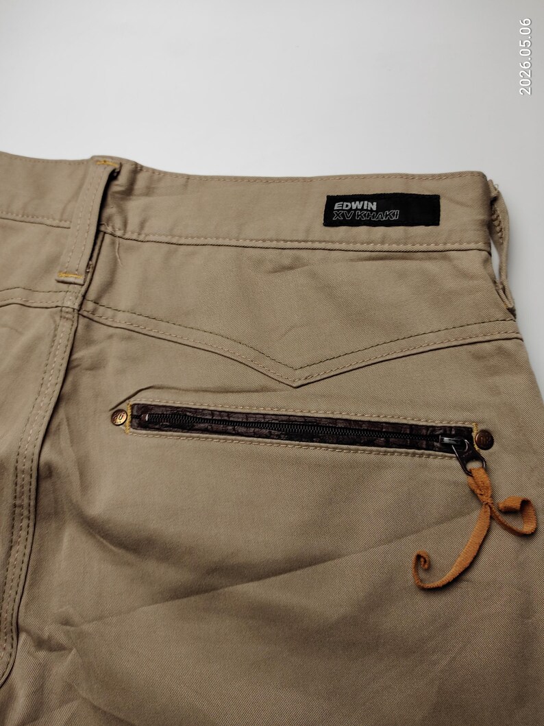 May include: Close-up of khaki-colored pants with a zipper pocket and a leather pull tab. The pants have a black label that reads "EDWIN XV KHAKI". The stitching is visible, and the overall design is casual.