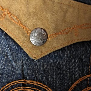 May include: Close-up of denim jeans with a tan suede pocket and a silver button. The pocket is outlined with orange stitching, and the button has a sunburst design. The jeans have a classic blue wash.