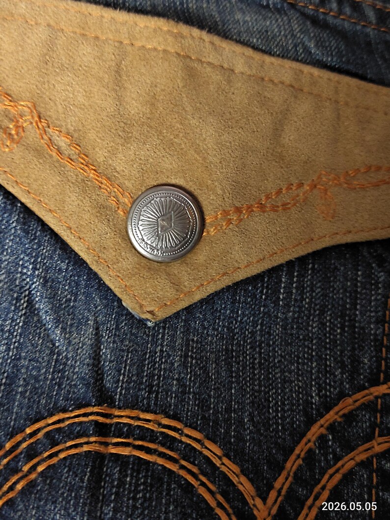 May include: Close-up of denim jeans with a tan suede pocket and a silver button. The pocket is outlined with orange stitching, and the button has a sunburst design. The jeans have a classic blue wash.