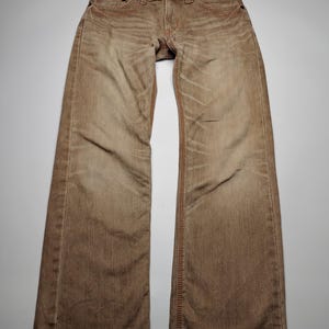 May include: Brown denim jeans with a faded appearance. The jeans have a classic five-pocket design, a button closure, and orange stitching. The jeans are laid flat on a white surface.