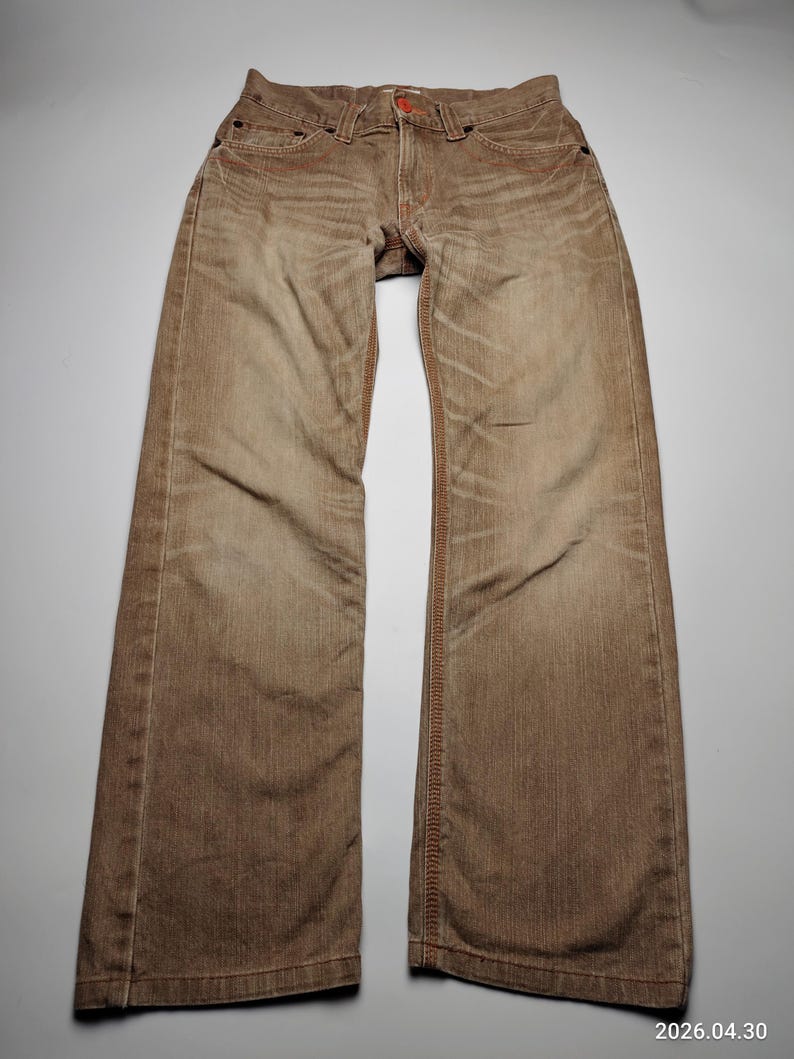 May include: Brown denim jeans with a faded appearance. The jeans have a classic five-pocket design, a button closure, and orange stitching. The jeans are laid flat on a white surface.