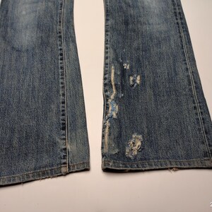 Peut inclure: Gros plan sur un jean en denim bleu avec une coupe &eacute;vas&eacute;e. Le jean pr&eacute;sente des d&eacute;tails us&eacute;s, notamment des d&eacute;chirures et des bords effiloch&eacute;s sur une jambe. Le denim a un aspect d&eacute;lav&eacute;.
