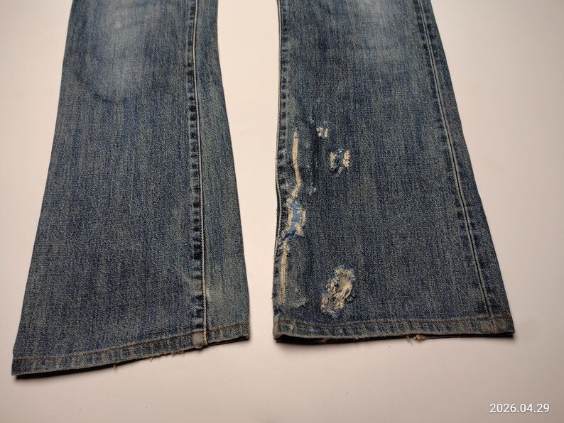 Peut inclure: Gros plan sur un jean en denim bleu avec une coupe &eacute;vas&eacute;e. Le jean pr&eacute;sente des d&eacute;tails us&eacute;s, notamment des d&eacute;chirures et des bords effiloch&eacute;s sur une jambe. Le denim a un aspect d&eacute;lav&eacute;.