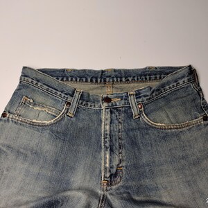 May include: Close-up of light blue denim jeans. The jeans have a classic five-pocket design, a button closure, and belt loops. The denim has a faded, worn appearance, with visible stitching and rivets. The jeans are laid flat against a white background.