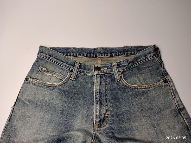 May include: Close-up of light blue denim jeans. The jeans have a classic five-pocket design, a button closure, and belt loops. The denim has a faded, worn appearance, with visible stitching and rivets. The jeans are laid flat against a white background.