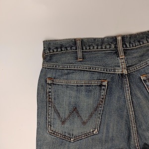 Peut inclure: Gros plan sur un jean en denim bleu avec une poche arri&egrave;re orn&eacute;e du logo Wrangler. Le jean pr&eacute;sente une conception classique &agrave; cinq poches et un d&eacute;lavage d&eacute;color&eacute;, montrant des signes d'usure. Les coutures sont visibles en couleur marron contrastante.
