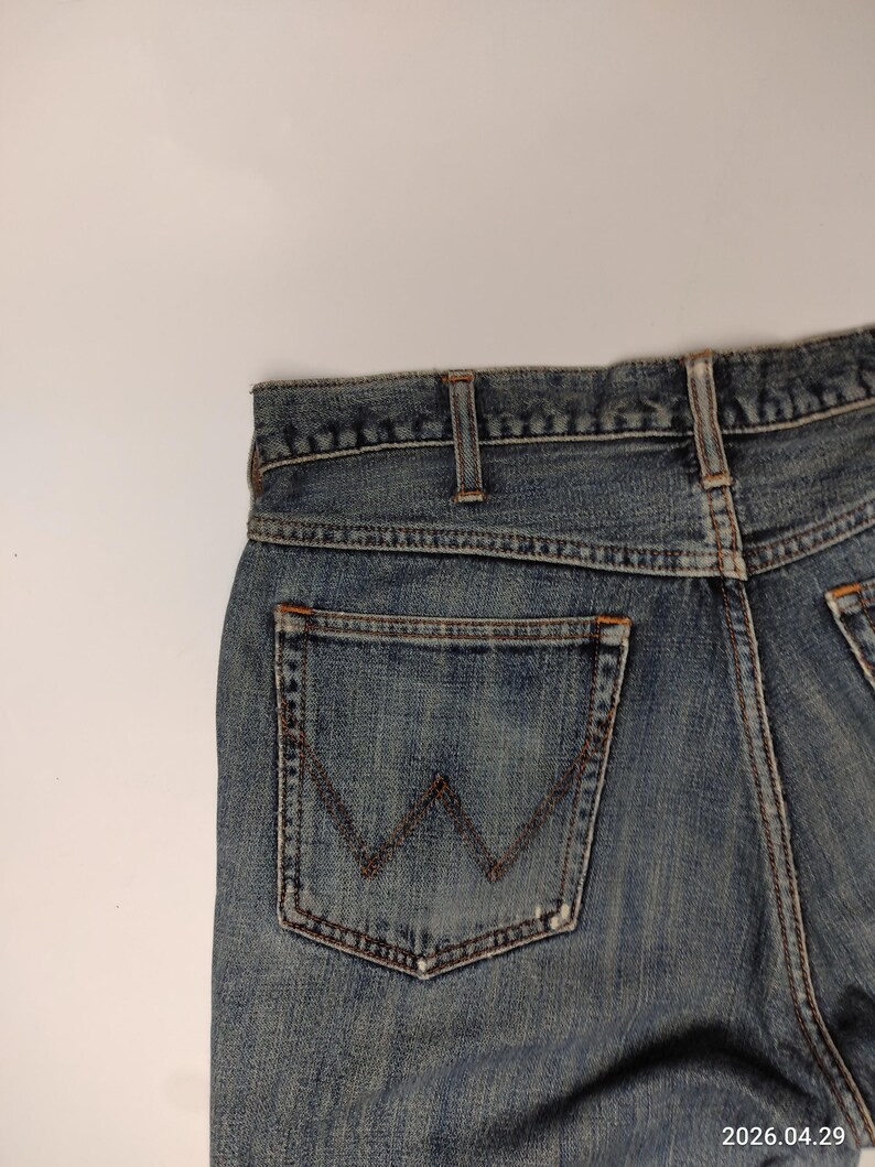 Peut inclure: Gros plan sur un jean en denim bleu avec une poche arri&egrave;re orn&eacute;e du logo Wrangler. Le jean pr&eacute;sente une conception classique &agrave; cinq poches et un d&eacute;lavage d&eacute;color&eacute;, montrant des signes d'usure. Les coutures sont visibles en couleur marron contrastante.