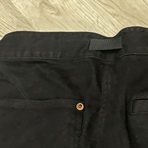 Puede incluir: Primer plano de la parte trasera de unos pantalones vaqueros negros. Los pantalones tienen una cintura con trabilla, un bolsillo trasero y un cierre de bot&oacute;n. El tejido vaquero es de color oscuro liso.
