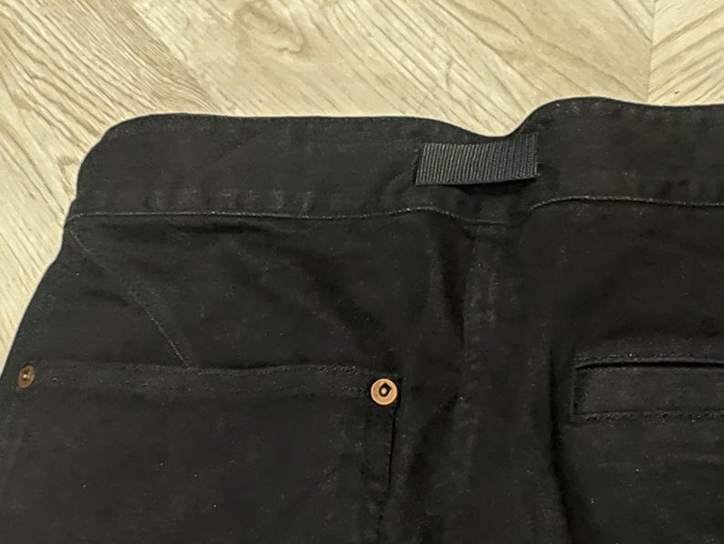 Puede incluir: Primer plano de la parte trasera de unos pantalones vaqueros negros. Los pantalones tienen una cintura con trabilla, un bolsillo trasero y un cierre de bot&oacute;n. El tejido vaquero es de color oscuro liso.