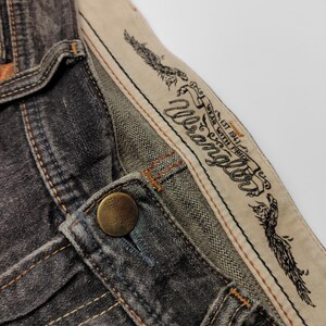 May include: Close-up of a pair of dark gray Wrangler jeans. The jeans feature a button closure, visible stitching, and a branded waistband with an eagle design and the text "Wrangler". The fabric appears worn.