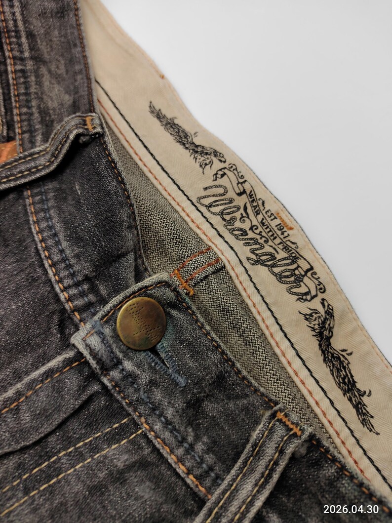 May include: Close-up of a pair of dark gray Wrangler jeans. The jeans feature a button closure, visible stitching, and a branded waistband with an eagle design and the text "Wrangler". The fabric appears worn.