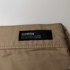 May include: Close-up of khaki-colored fabric with a black label. The label reads "EDWIN XV KHAKI" in white lettering. The fabric has visible stitching along the top edge. The overall tone is neutral, suggesting a garment.