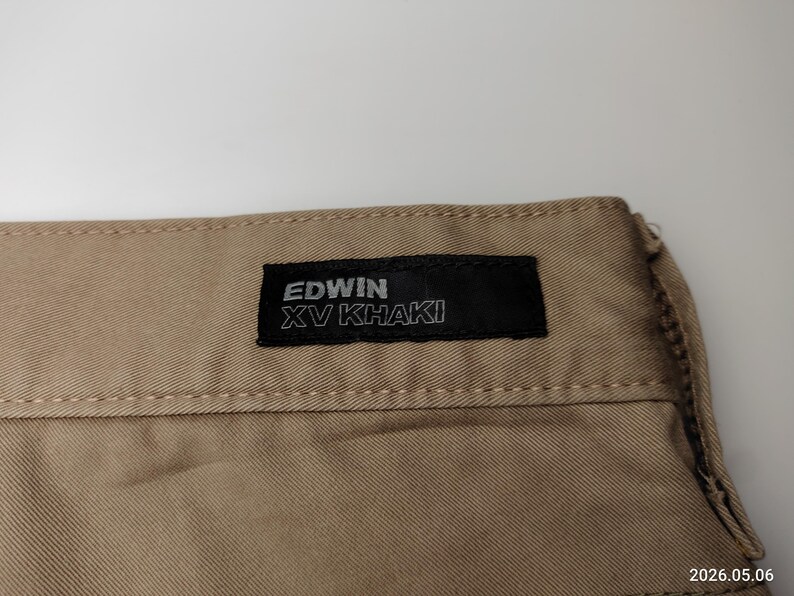 May include: Close-up of khaki-colored fabric with a black label. The label reads "EDWIN XV KHAKI" in white lettering. The fabric has visible stitching along the top edge. The overall tone is neutral, suggesting a garment.