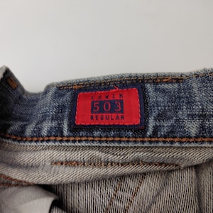 May include: Close-up of denim jeans with a red tag. The tag reads "EDWIN 503 REGULAR" in white lettering. The jeans are a faded blue color with visible stitching and a worn appearance, suggesting a vintage or well-loved item.