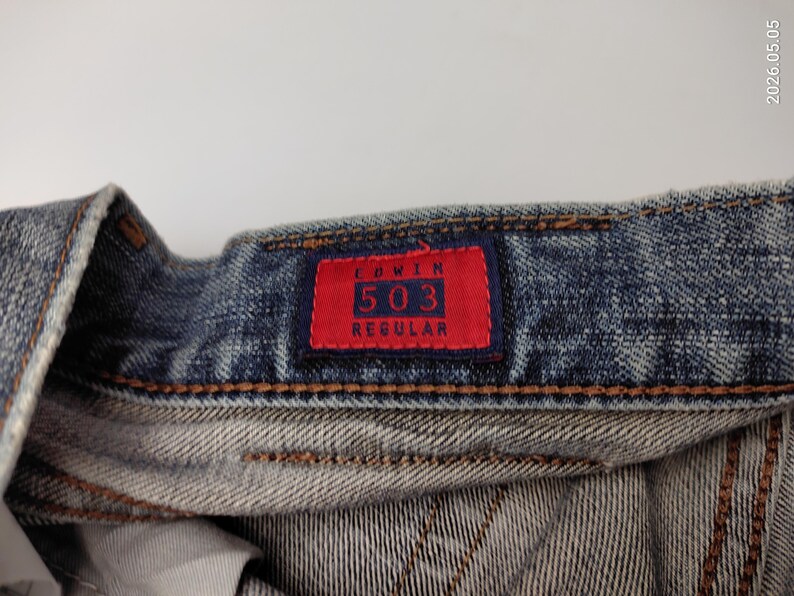 May include: Close-up of denim jeans with a red tag. The tag reads "EDWIN 503 REGULAR" in white lettering. The jeans are a faded blue color with visible stitching and a worn appearance, suggesting a vintage or well-loved item.