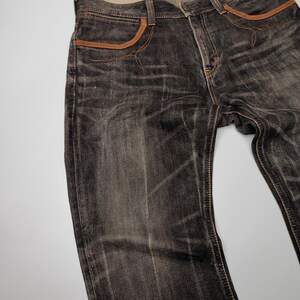 May include: Dark gray denim jeans with brown pocket trim and stitching. The jeans have a faded, worn look. The waistline has a light-colored interior with printed text.