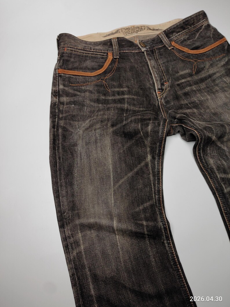 May include: Dark gray denim jeans with brown pocket trim and stitching. The jeans have a faded, worn look. The waistline has a light-colored interior with printed text.