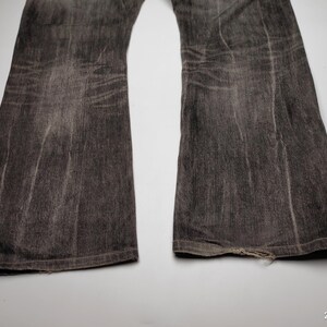 May include: Close-up of a pair of faded black denim jeans with a flared leg. The jeans show signs of wear, with visible fading and distressing. The bottom hem of one leg is slightly frayed.