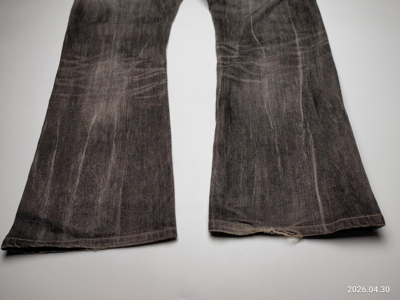 May include: Close-up of a pair of faded black denim jeans with a flared leg. The jeans show signs of wear, with visible fading and distressing. The bottom hem of one leg is slightly frayed.