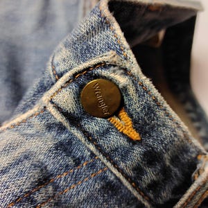 Peut inclure: Gros plan sur le col d'une veste en jean bleu avec un bouton en laiton. Le bouton porte le nom de la marque "Wrangler". Le denim pr&eacute;sente des coutures orange visibles et un aspect d&eacute;lav&eacute;.