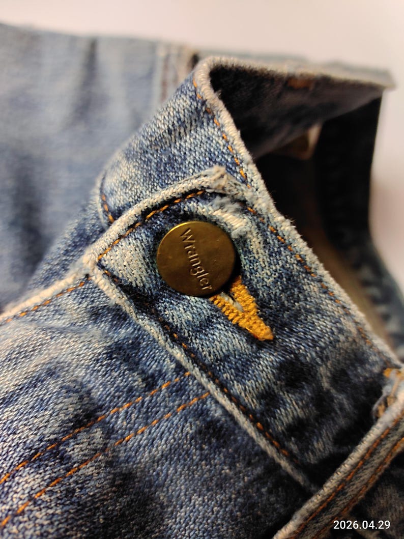 Peut inclure: Gros plan sur le col d'une veste en jean bleu avec un bouton en laiton. Le bouton porte le nom de la marque "Wrangler". Le denim pr&eacute;sente des coutures orange visibles et un aspect d&eacute;lav&eacute;.