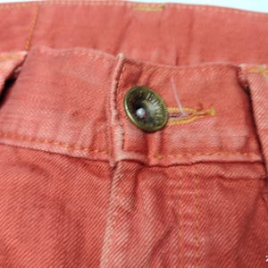 May include: Close-up of coral-colored denim pants. The image highlights the button closure, belt loops, and stitching details. The button is a tarnished brass color. The fabric has a textured appearance.