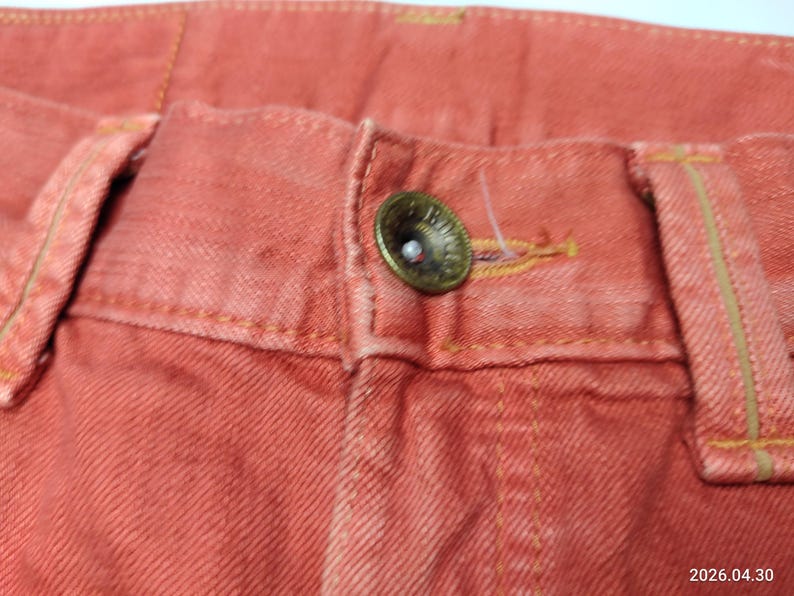 May include: Close-up of coral-colored denim pants. The image highlights the button closure, belt loops, and stitching details. The button is a tarnished brass color. The fabric has a textured appearance.