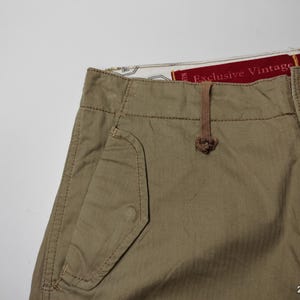 May include: Close-up of khaki-colored pants with a pocket detail and a button closure. The pants have a drawstring and a tag that reads "Exclusive Vintage". The fabric appears to be a durable cotton blend.