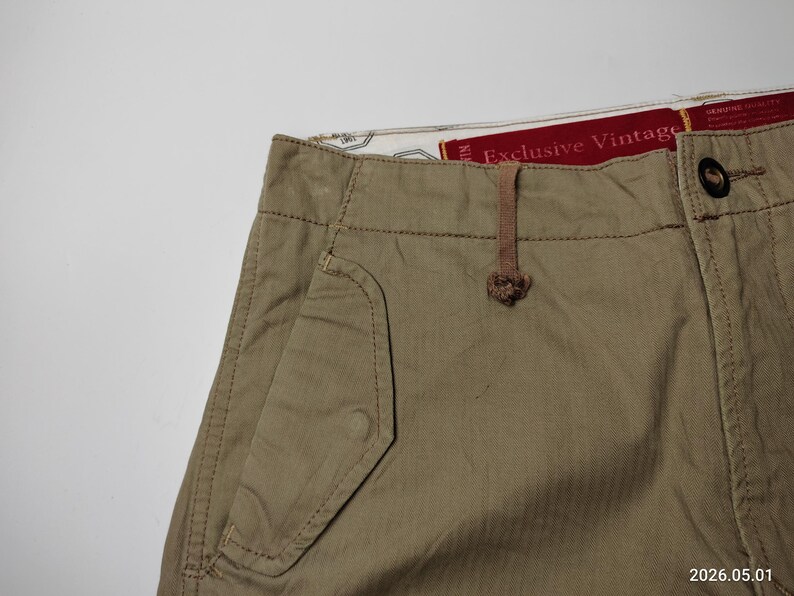 May include: Close-up of khaki-colored pants with a pocket detail and a button closure. The pants have a drawstring and a tag that reads "Exclusive Vintage". The fabric appears to be a durable cotton blend.