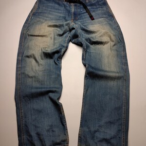 Blue denim pants with a black belt. The pants have a relaxed fit and a faded wash, with visible wear patterns. The belt has a red accent. The pants are laid flat on a white surface.