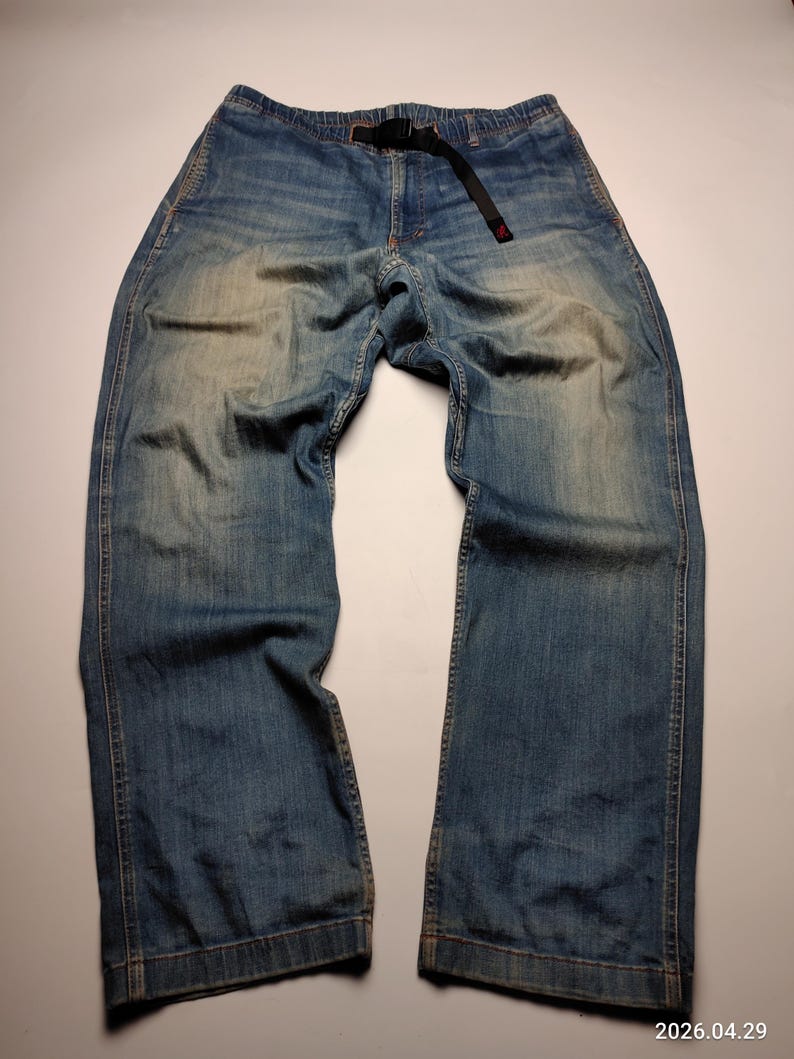 Blue denim pants with a black belt. The pants have a relaxed fit and a faded wash, with visible wear patterns. The belt has a red accent. The pants are laid flat on a white surface.