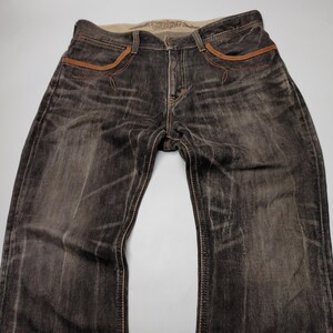 May include: Dark wash denim jeans with brown stitching and accents. The jeans have a faded, worn look, with brown leather-like trim around the pockets. The waistband is a lighter color, and the jeans appear to be new.