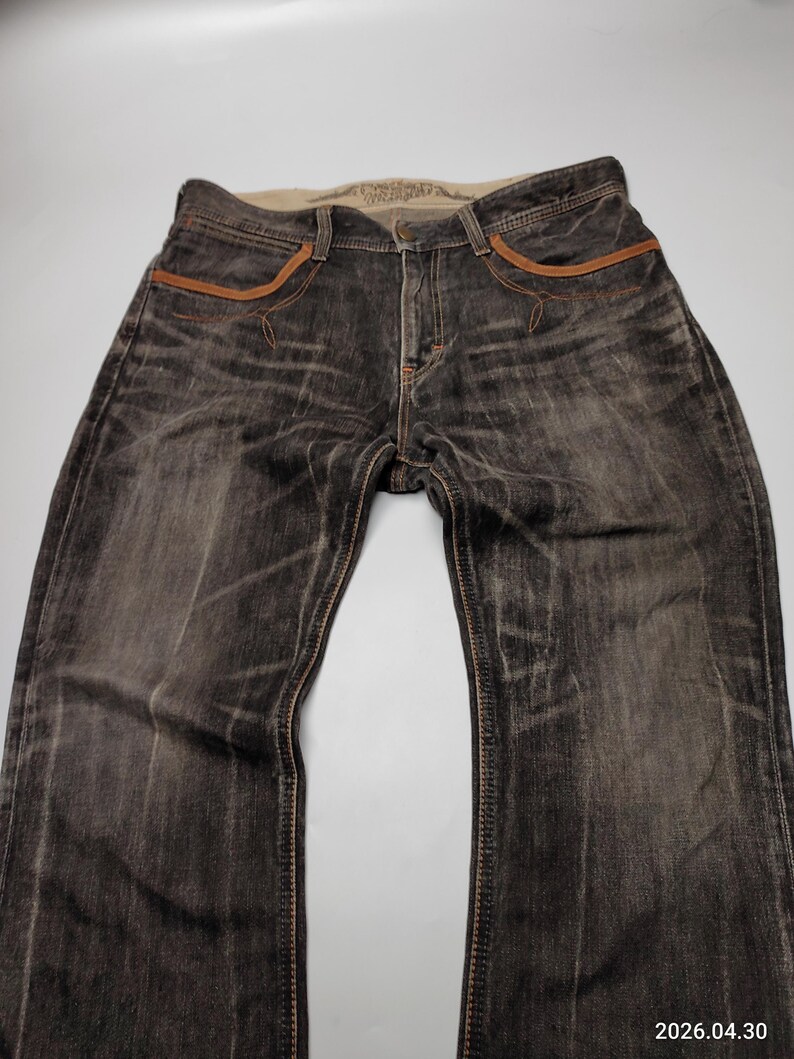May include: Dark wash denim jeans with brown stitching and accents. The jeans have a faded, worn look, with brown leather-like trim around the pockets. The waistband is a lighter color, and the jeans appear to be new.