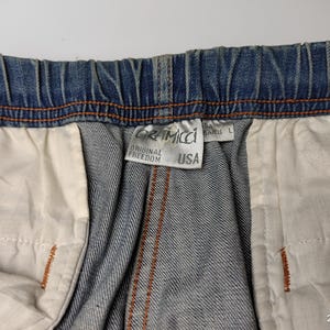 May include: Close-up of blue denim shorts with an elasticated waistband and orange stitching. The shorts have a white interior lining. A tag reads "Gramicci Original Freedom USA".