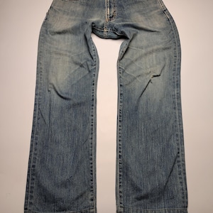 May include: Light blue denim jeans with a faded wash and a distressed look. The jeans have a classic five-pocket design and a button and zipper closure. There is a tear on the right leg. The jeans are laid flat on a white surface.