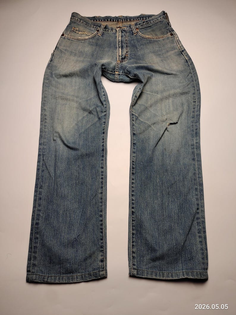 May include: Light blue denim jeans with a faded wash and a distressed look. The jeans have a classic five-pocket design and a button and zipper closure. There is a tear on the right leg. The jeans are laid flat on a white surface.