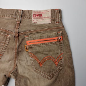 May include: Close-up of brown denim jeans with orange stitching and zipper details. The back pocket features a distinctive orange embroidered design. The Edwin brand label is visible at the waistband.
