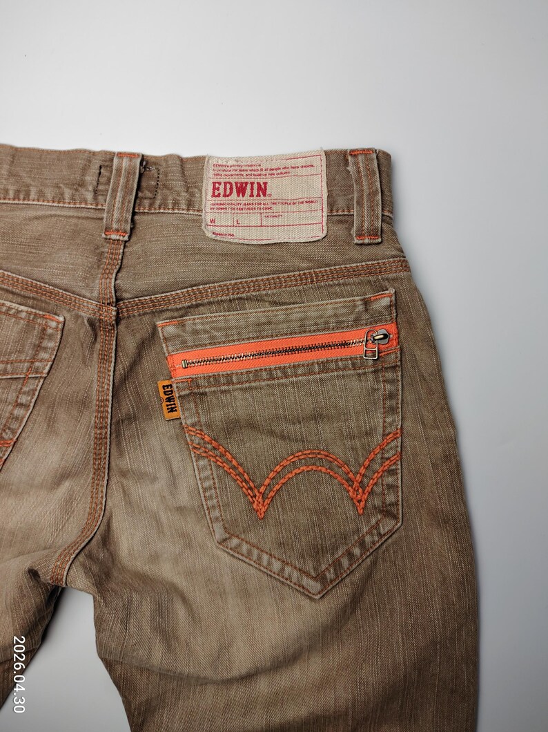 May include: Close-up of brown denim jeans with orange stitching and zipper details. The back pocket features a distinctive orange embroidered design. The Edwin brand label is visible at the waistband.