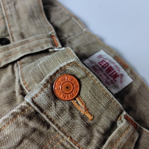 May include: Close-up of tan-colored denim pants with orange stitching and a matching button. The button is embossed with the brand name "EDWIN". A small tag with the brand name is also visible.