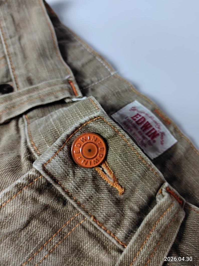 May include: Close-up of tan-colored denim pants with orange stitching and a matching button. The button is embossed with the brand name "EDWIN". A small tag with the brand name is also visible.