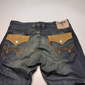 May include: Blue denim jeans with tan suede-like pocket details and orange stitching. The back pockets feature a distinctive design with a silver button closure. The Edwin brand label is visible at the waistband.